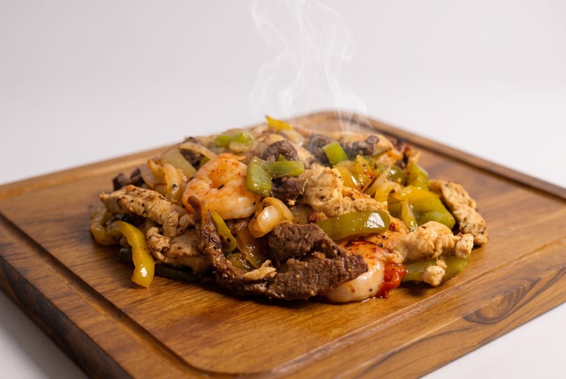 Steaming fajitas with shrimp, chicken, beef, onions, and bell peppers on a wooden board.