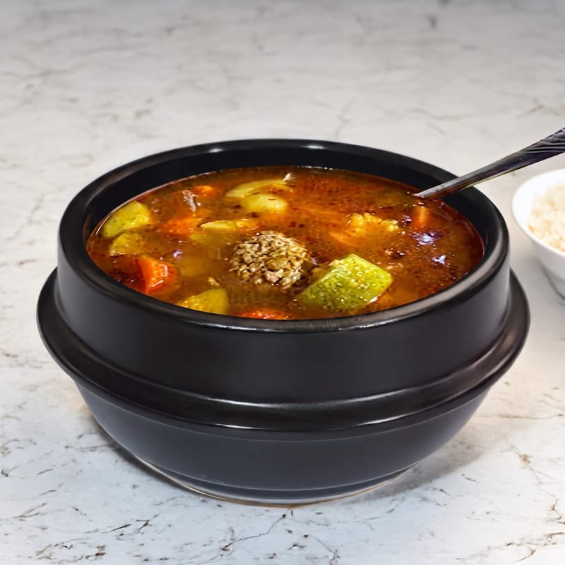 A black bowl of Korean stew with vegetables and a meatball, with a spoon and rice.