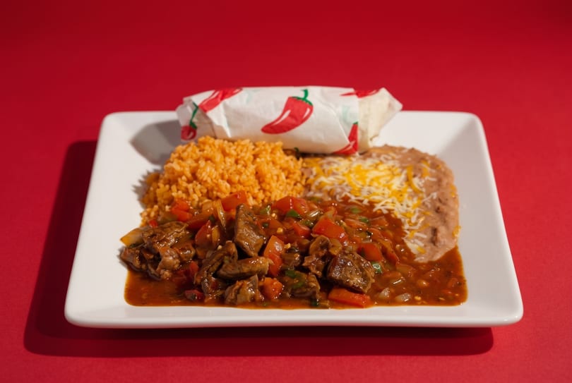 A plate of carne guisada with rice, refried beans, and a burrito on a red background.