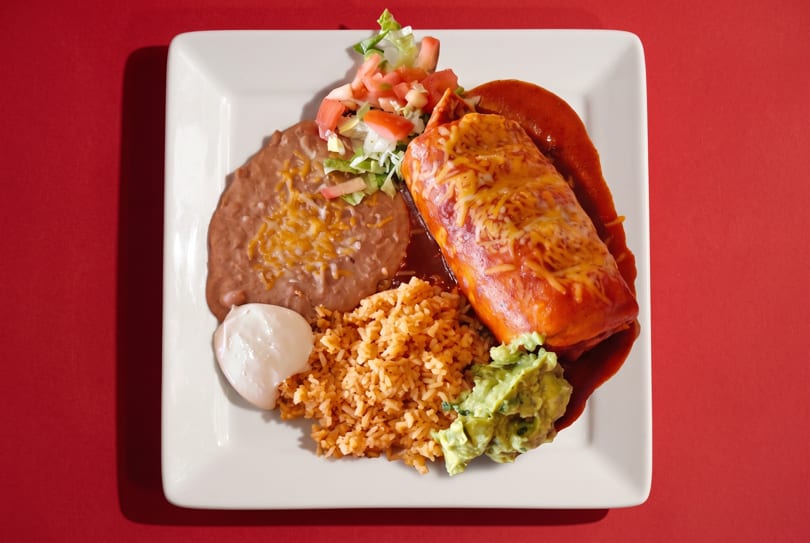 A cheese-covered burrito with red sauce, rice, refried beans, guacamole, sour cream, and salad.